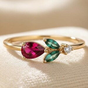 May include: A gold ring featuring a pear-shaped, deep red gemstone and two green, leaf-shaped stones. A small, round, clear stone is set between the green stones. The ring is set on a cream-colored surface.