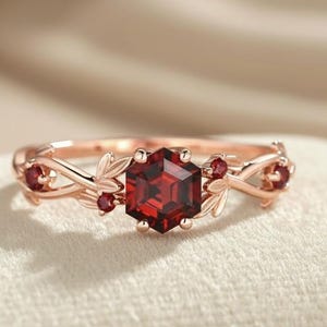 May include: Rose gold ring featuring a hexagonal, deep red gemstone as the centerpiece. The ring is adorned with smaller red gemstones and delicate leaf and vine detailing, creating an elegant, nature-inspired design. The ring is set on a neutral-colored surface.