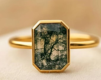 May include: A gold ring featuring a rectangular moss agate stone. The stone has a green and white pattern resembling a landscape. The ring's band and bezel are gold-colored, creating a classic and elegant design.