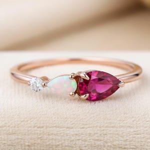 Peut inclure: Une bague en or rose ornée de trois pierres précieuses : un petit diamant rond, une opale en forme de poire et un rubis en forme de poire plus grand. La bague est posée sur un fond neutre.