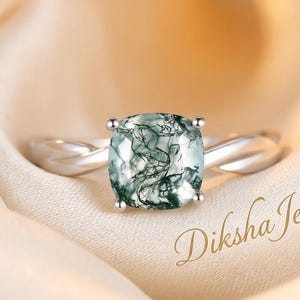 May include: A silver ring featuring a cushion-cut moss agate gemstone with dark green dendritic inclusions. The band has a twisted metal design. The text Diksha Jewel is visible in the lower right corner of the image.