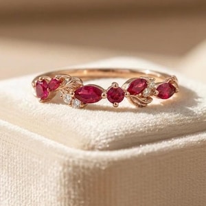 May include: A rose gold ring featuring a series of marquise and round-cut red rubies interspersed with small clear stones and delicate metal leaf motifs. The band has a polished finish and follows a curved, organic arrangement of gemstones.