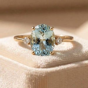 Può includere: Un anello in oro con una grande pietra preziosa azzurra taglio ovale al centro, affiancata da due pietre bianche trasparenti più piccole taglio rotondo su entrambi i lati. La fascia è sottile e lucida con una classica montatura a griffe.