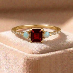 May include: A gold ring featuring a central square-cut deep red garnet gemstone flanked by two small round white opals. The thin gold band is accented with tiny pavé-set clear stones along the sides, creating a delicate and refined appearance.