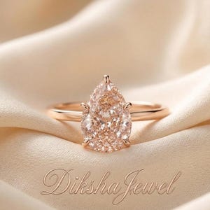 Op de afbeelding: Een ring van roségoud met een peervormige, perzikkleurige edelsteen die door zes pootjes op een gladde band wordt vastgehouden. De tekst Diksha Jewel is in cursief schrift zichtbaar onderaan het beeld, gecentreerd onder de ring.