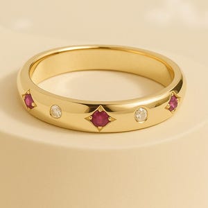 Pode incluir: Um anel de banda de ouro com detalhes alternados de rubis e diamantes. O anel tem um acabamento polido e um design clássico. Os rubis são engastados em forma de diamante e os diamantes são redondos. Uma joia para uso diário.