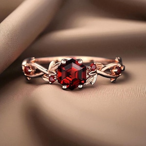 Könnte beinhalten: Ein roségoldener Ring mit einem großen, facettierten, sechseckigen roten Edelstein. Der Ring hat zarte Blatt- und Beerenakzente am Band, die ein elegantes, von der Natur inspiriertes Design hinzufügen. Der Ring ist vor einem neutralen Hintergrund platziert.