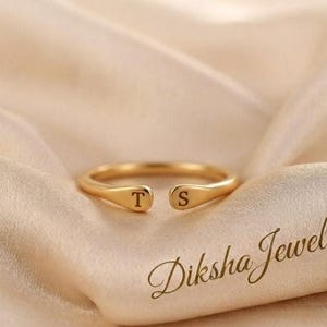 Puede incluir: Un anillo abierto de tono dorado con dos extremos planos y redondeados, cada uno grabado con una letra mayúscula, una 'T' a la izquierda y una 'S' a la derecha. El anillo tiene un acabado liso y pulido con un diseño de banda simple.