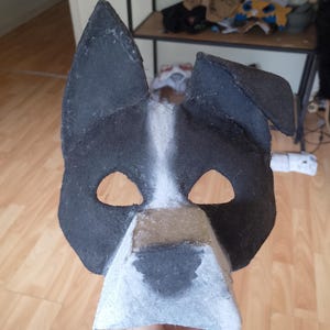May include: A handmade dog mask in black and white. The mask has large, pointed ears, eye holes, and a snout. It's made from felt or a similar material, designed to be worn on the face. A fun costume accessory.