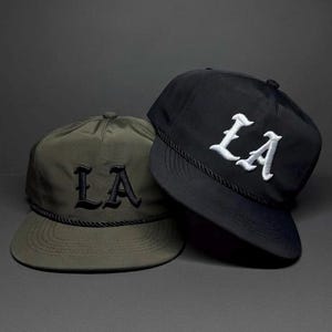 Może przedstawiać: Dwie czapki bejsbolowe, jedna oliwkowa i jedna czarna, są prezentowane na ciemnoszarym tle. Obie czapki mają wyszyte litery "LA" w stylizowanej czcionce. Czarny sznur zdobi rondo każdej czapki.