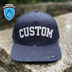 Op de afbeelding: Een marineblauwe, waterbestendige baseballcap met het woord "CUSTOM" in witte blokletters. De cap is nat met waterdruppels en een "WATER RESISTANT" logo is zichtbaar in de linkerbovenhoek.