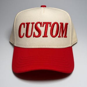 Könnte beinhalten: Eine Baseballkappe mit cremefarbener Krone und rotem Schirm. Das Wort "CUSTOM" ist rot auf die Vorderseite gestickt. Die Kappe hat einen Knopf oben und einen gebogenen Schirm.