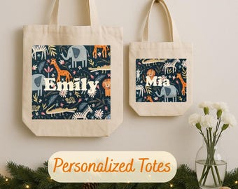 May include: Two canvas tote bags, one large and one small, hanging against a white wall. Each bag features a rectangular print with a dark blue background and colorful animal illustrations. The larger bag says "Emily" and the smaller bag says "Mia". The text "Personalized Totes" is below.