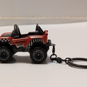 May include: A red and black toy jeep keyring. The miniature vehicle has black wheels, a black roll bar, and a checkered flag design on the front. The keyring is black.