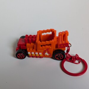 May include: A toy car keychain in orange and red. The car is made from small, pixelated blocks, with black wheels and red detailing. It has a red keyring attached. The car features a flame design on its side.