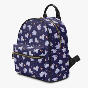 Puede incluir: Una mochila azul marino con un estampado repetido de conejos alados, lunas y estrellas. La mochila tiene cremalleras doradas, un asa negra y un pequeño bolsillo delantero. El diseño es caprichoso y adecuado para el uso diario.