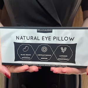 May include: A rectangular package labeled "NATURAL EYE PILLOW" is held in front of a person. The package features text and icons for black beans, chrysanthemum, and lavender. The text on the package also includes "RELAXALIGNS". The package is white with black text and graphics.