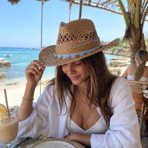 May include: A straw cowboy hat with a light blue band and seashell accents. The hat has a perforated crown and a wide brim. The hat is worn by a person wearing a white shirt, sitting at a table with a drink.