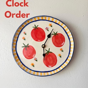 May include: A round wall clock with a cream-colored face featuring four hand-painted red tomatoes and small orange and purple flowers. The clock has a yellow and white striped border with a blue rim and black hands. Text reads Custom Clock Order.