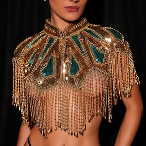 May include: A gold and teal beaded cape with fringe. The cape features a high collar and geometric teal accents. Long gold beaded fringe hangs from the bottom of the cape. The garment is embellished with sequins and beads.