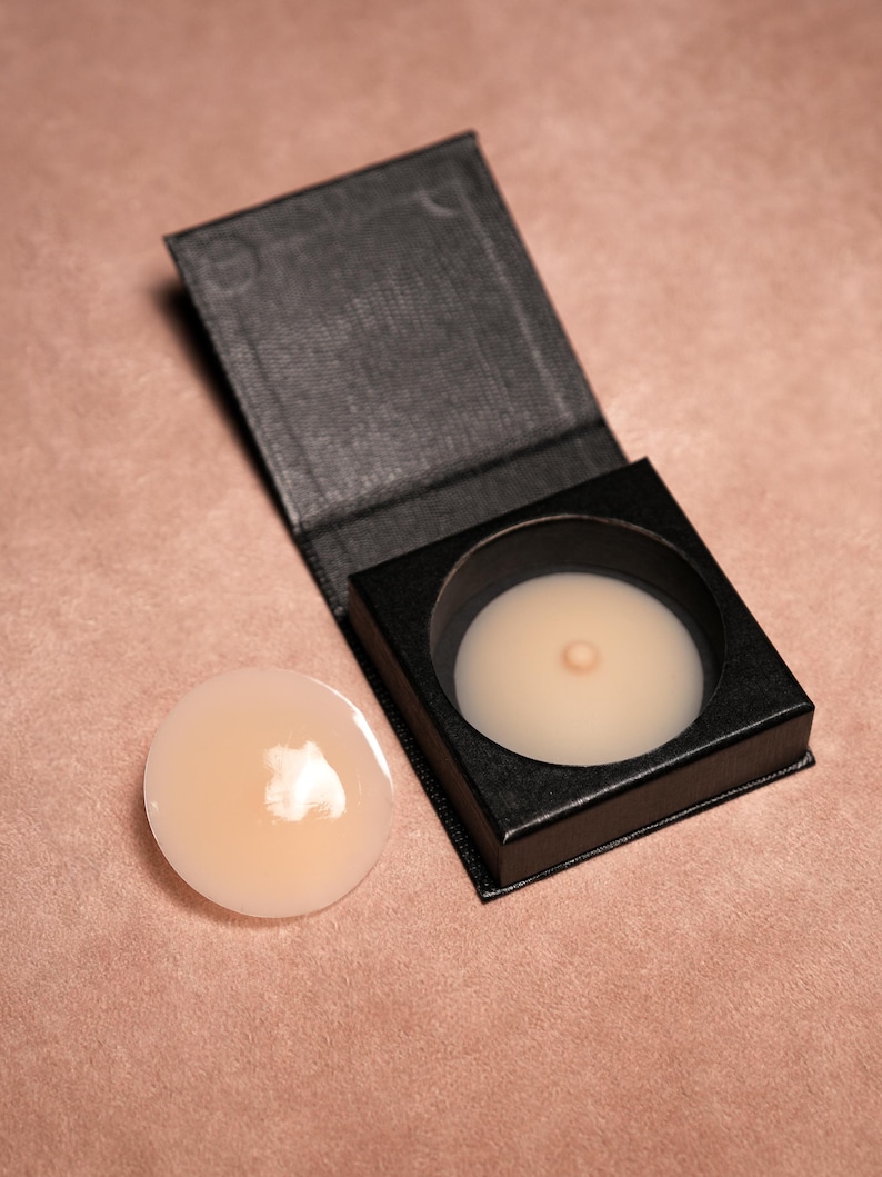May include: A pair of flesh-toned silicone nipple covers, one inside a black box and the other lying next to it. The covers are round with a central raised area. The box is open, revealing the product.