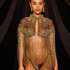 May include: A gold beaded cape with a high collar and fringe detailing. The sheer fabric is adorned with intricate beadwork, creating a glamorous and eye-catching design. The cape is a statement piece for special occasions.