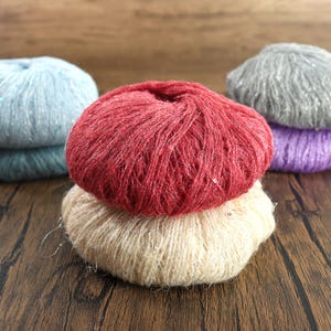 May include: Several balls of yarn in shades of red, cream, blue, grey, and purple. The yarn has a soft, textured appearance with a subtle sheen. The balls are arranged on a wooden surface, suggesting a craft or hobby theme.