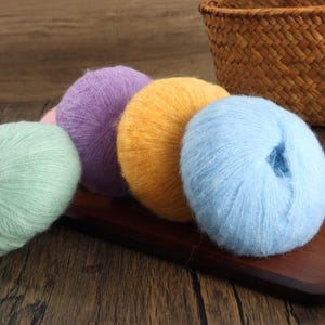 Könnte beinhalten: Eine Sammlung bunter Wollknäuel in den Farben Hellgrün, Lila, Gelb und Blau. Die Wollknäuel sind auf einem dunklen Holztablett angeordnet, im Hintergrund ein geflochtener Korb. Die Wolle wirkt weich und flauschig.