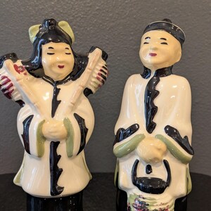 May include: A pair of vintage ceramic figurines depicting Asian figures. The figurines are dressed in white robes with black and green accents. One figure holds brushes, the other a basket. The bases are black.
