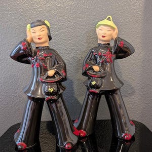 May include: Two ceramic figurines of people in traditional Chinese attire. The figures are dressed in black outfits with red floral accents and hold small black purses. The figurines have a glossy finish and are posed with one hand near their head.