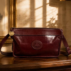 May include: A dark burgundy leather crossbody bag with a rectangular shape, a top zipper closure, and an adjustable shoulder strap. The front features a circular embossed logo patch, and a gold-toned metal charm is attached to the zipper pull.