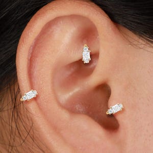 May include: Close-up of an ear adorned with three gold-colored earrings. Each earring features a cluster of small, sparkling white gemstones. The earrings are set in a delicate, minimalist design, adding a touch of elegance.