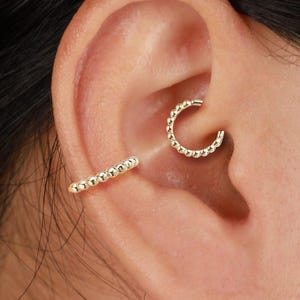 May include: A close-up shot of an ear featuring two pieces of gold jewellery. One is a small, beaded hoop earring, and the other is a beaded bar earring. The jewellery is set against the skin of the ear.