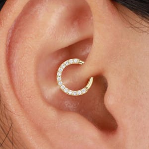 Puede incluir: Un pendiente de aro dorado con bisagras y pequeñas piedras preciosas transparentes engastadas a lo largo de la curva exterior. El pendiente se lleva en el cartílago de la oreja, mostrando un diseño delicado y brillante. Joyería.