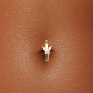 May include: A gold belly button ring with a butterfly design. The butterfly has white accents and is centered on the ring. The ring is shown on a person's abdomen, with blue denim jeans visible at the bottom of the image.