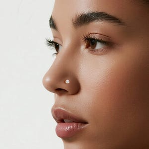 Könnte beinhalten: Seitenansicht einer Person mit Nasenpiercing und zwei kleinen Ohrringen. Der Schmuck ist klein und rund, wahrscheinlich aus reflektierendem Material. Die Person hat dunkles Haar, das zurückgebunden ist, und trägt ein schwarzes Oberteil.