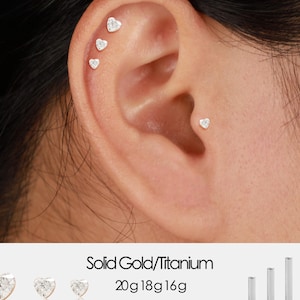 Puede incluir: Primer plano de una oreja con múltiples pendientes de botón en forma de corazón. Los pendientes son pequeños y parecen estar hechos de un metal brillante. El texto en la parte inferior dice "Solid Gold/Titanium" y enumera tamaños de 5 mm a 10 mm.