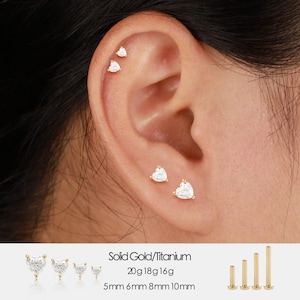 May include: Ear piercings with heart-shaped, clear gemstones set in gold. The image shows multiple earrings of varying sizes, with the text "Solid Gold/Titanium" and size specifications: 20g 18g 16g, 5mm 6mm 8mm 10mm.