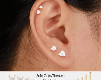May include: Ear piercings featuring heart-shaped, clear gemstones set in gold. The image shows multiple earrings of varying sizes, with the text "Solid Gold/Titanium" and size specifications: 20g 18g 16g, 5mm 6mm 8mm 10mm.