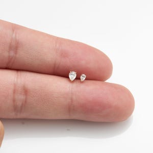 Puede incluir: Dos pendientes peque&ntilde;os en forma de l&aacute;grima con piedras claras y brillantes. Los pendientes est&aacute;n engarzados en metal dorado y se muestran sobre un fondo claro. Son una joya delicada.