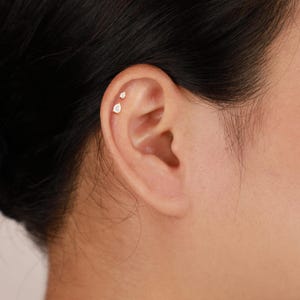 Puede incluir: Primer plano de una oreja con dos pendientes peque&ntilde;os de color dorado. El pendiente superior es un peque&ntilde;o y redondo, mientras que el pendiente inferior tiene forma de l&aacute;grima. Ambos pendientes tienen una piedra transparente y brillante.