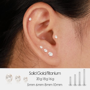 Puede incluir: Primer plano de una oreja adornada con múltiples pendientes de botón claros y brillantes. La imagen también muestra pendientes adicionales y el texto "Solid Gold/Titanium" con medidas de calibre y milímetros.