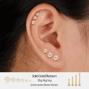 Puede incluir: Pendientes de oro y titanio con gemas transparentes se muestran en una oreja. Los pendientes son de varios tamaños, que van de 5 mm a 10 mm. El texto "Solid Gold/Titanium" y los tamaños son visibles.