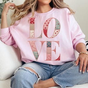 Puede incluir: Una sudadera rosa claro con la palabra "LOVE" en un diseño de patchwork. Las letras están decoradas con corazones y otros patrones en rojo, rosa y turquesa. La sudadera es de estilo casual.