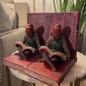 May include: A pair of bookends featuring two monkey figures reading books. The monkeys wear red hats and green vests. The bookends are designed to look like a large, open book with red pages and a distressed finish.