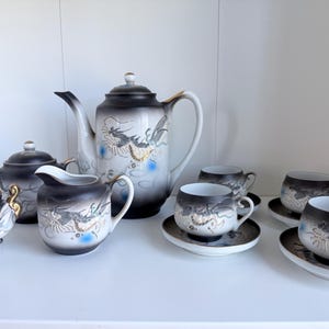 May include: A complete Japanese tea set with a black and white dragon design. The set includes a teapot, creamer, sugar bowl, and cups with saucers. The porcelain set has gold accents and is displayed on a white shelf.