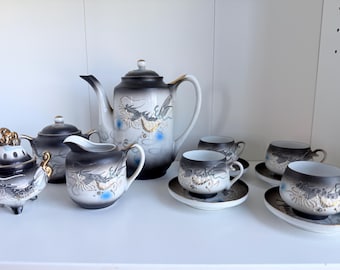May include: A complete Japanese tea set with a black and white dragon design. The set includes a teapot, creamer, sugar bowl, and cups with saucers. The porcelain set has gold accents and is displayed on a white shelf.