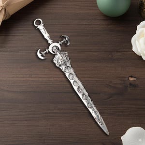 May include: A silver letter opener with a dagger design. The handle features a ring, crescent moons, and decorative carvings. The blade is adorned with circular embellishments and tapers to a point. The opener is displayed on a dark wooden surface.
