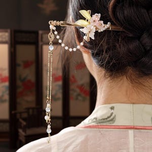 May include: A gold-toned hair stick with floral and pearl accents. The hair accessory features a cluster of pink and white flowers, a string of pearls, and dangling chains with iridescent beads. The hair stick is inserted into a dark bun.