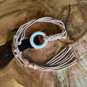 May include: A beige macrame plant hanger with a white ring at the top. The plant hanger is made of twisted rope and has a tassel at the bottom. The background is a wooden surface.